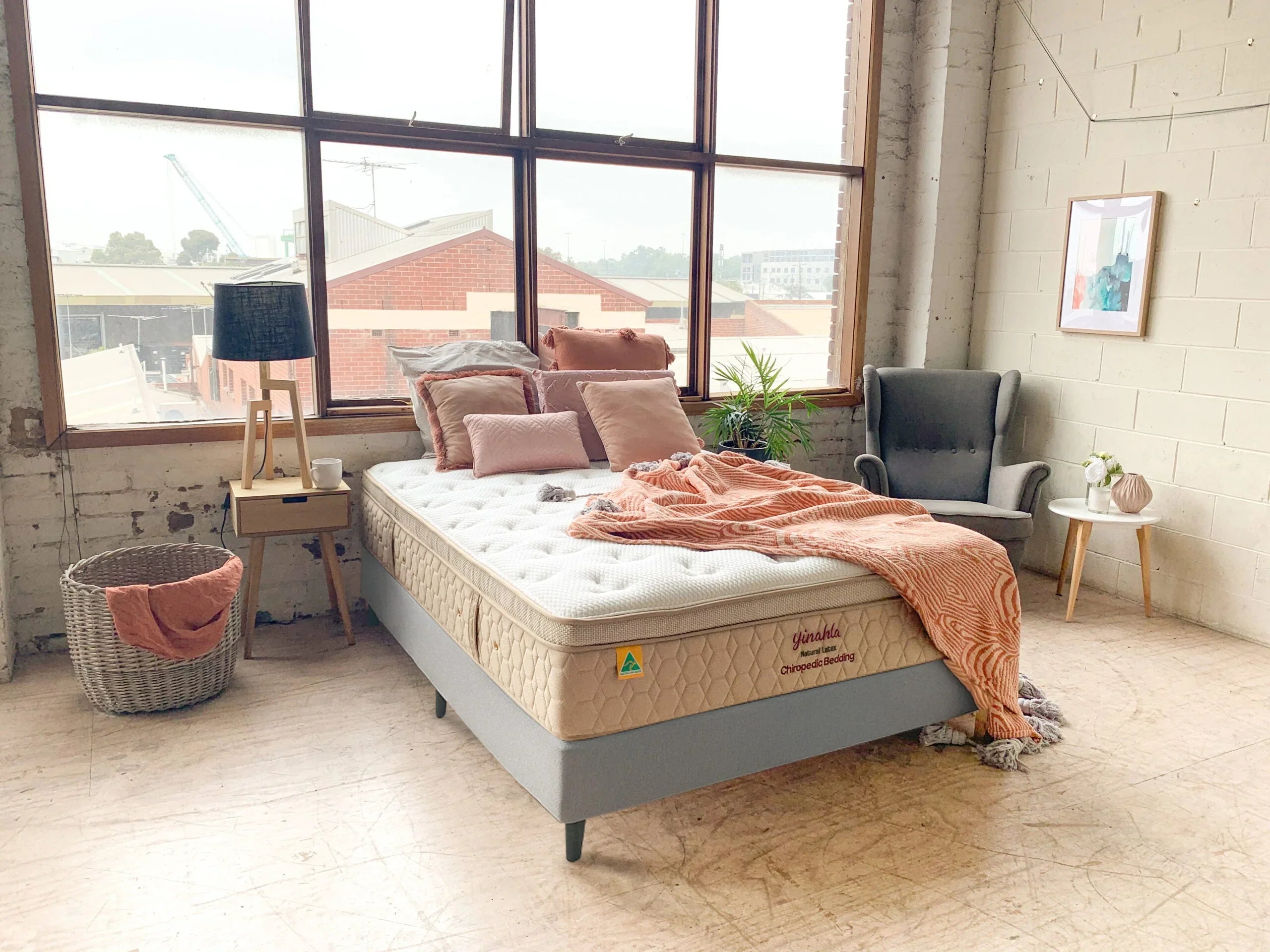 Natural latex mattress in a bright bedroom with pink pillows, blanket, grey chair, and large window