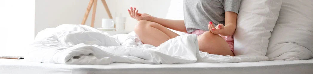 Woman meditating cross-legged on a white bed with cozy linens in a bright minimalist bedroom