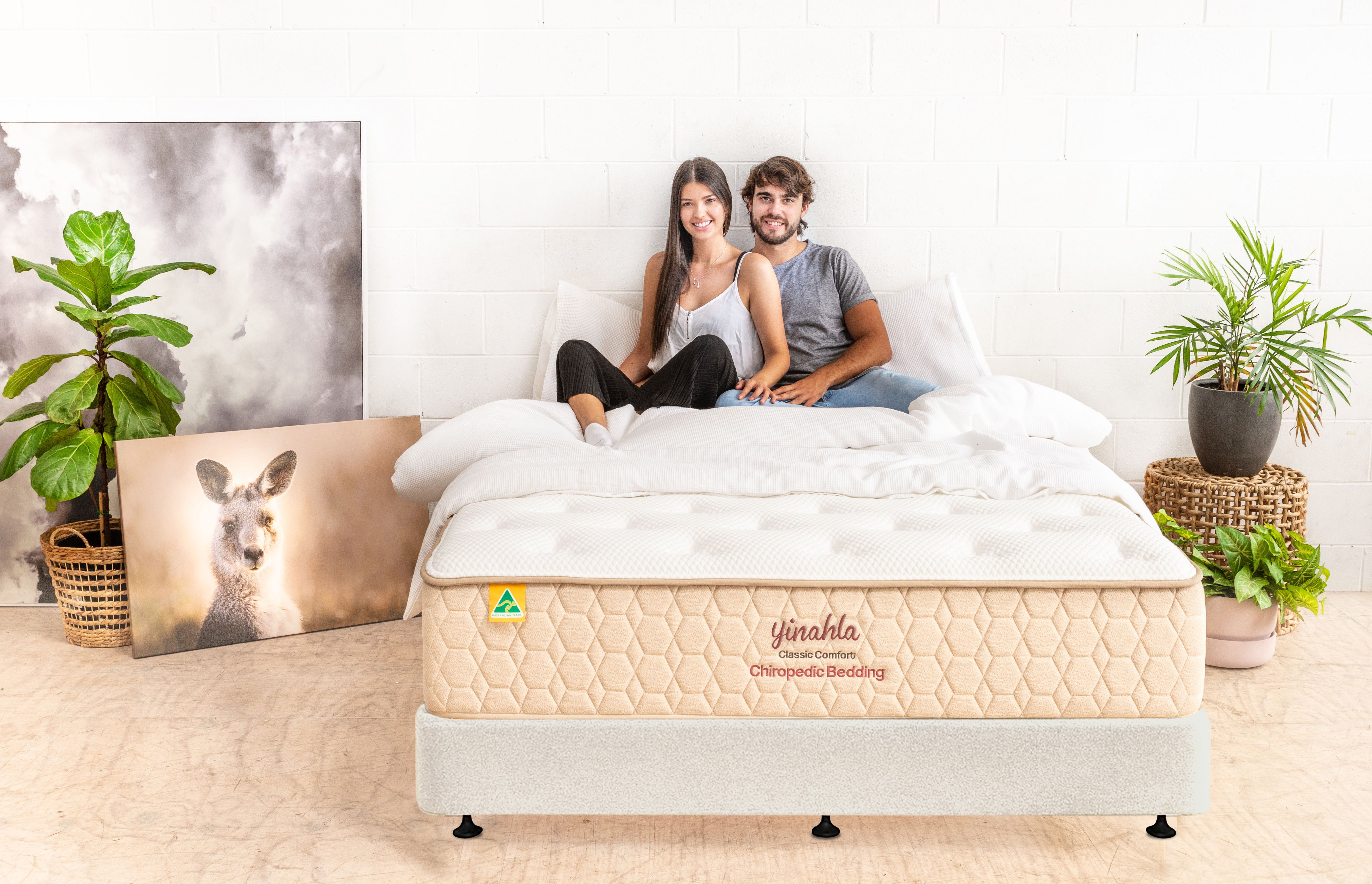 Young couple sitting on yinahla Classic Comfort Chiropedic mattress in bright, stylish bedroom with plants and kangaroo photo